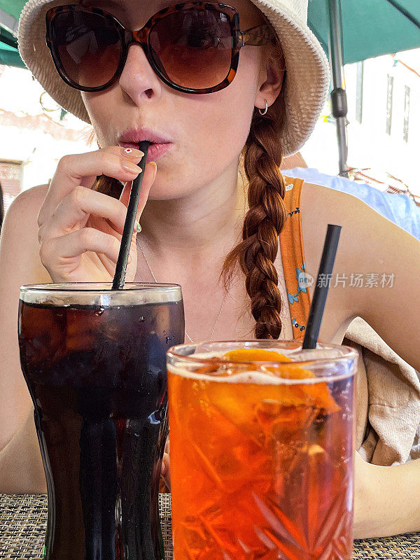 有魅力的红发女人用塑料吸管喝着可乐饮料和冰块，Spritz Veneziano鸡尾酒饮料和橙子片，在户外戴着太阳镜，遮阳帽和太阳裙，户外用餐设置背景